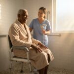 Senior seated on a sturdy shower chair in a bright accessible bathroom with grab bar and handheld shower, caregiver nearby